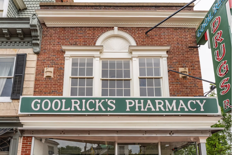A storefront with a green sign reading “GOOLRICK’S PHARMACY” in front of a red brick building, white trim, and a decorative arch above the windows.