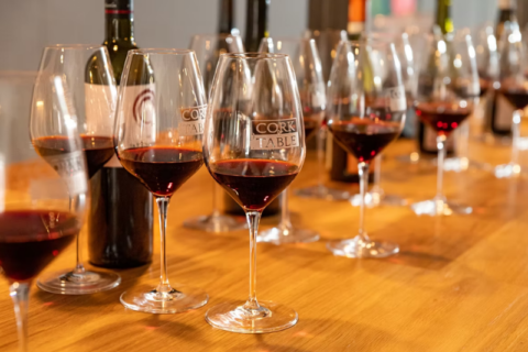 A row of wine glasses filled with red wine on a wooden table, with several bottles in the background and a blurred dining setting behind them.
