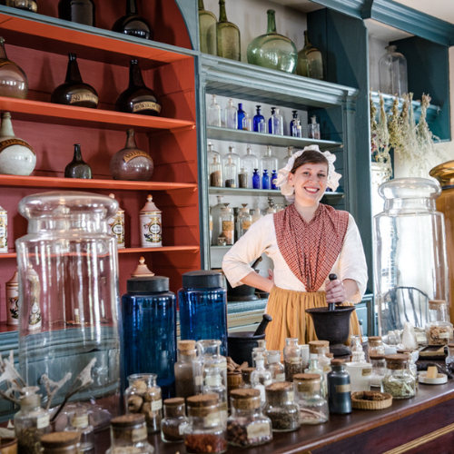 lady smiling at the camera behind all the medicinal herb bottles on the counter at the Hugh Mercer Apothecary