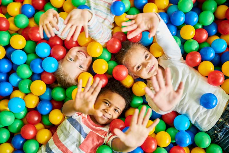 3 kids laying a in a ball pit. Kids are holding their hands up to the camera and smiling