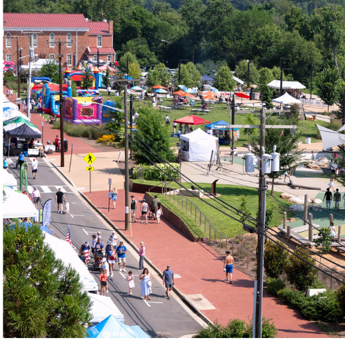 A bustling outdoor festival with inflatable bounce houses, tents, food vendors, and people strolling along a brick pathway and Sophia street.