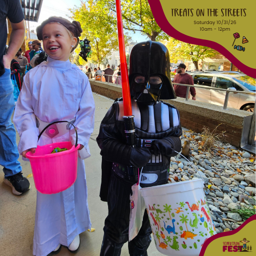 A child in a white costume holding a pink bucket of candy stands beside a person in black-and-white armor with a red saber on the front porch of the Visitor Center with autumn trees and rocks.
