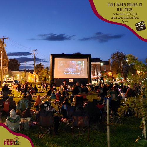 Outdoor movie at Riverfront Park with a large crowd seated on chairs and blankets in a park, warm streetlights lining the path, and a big illuminated movie screen stage under a deep blue sky.