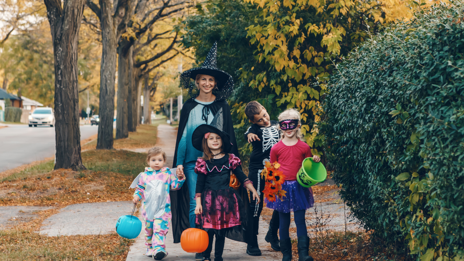 Trick or Treaters on the street. Lady as a witch and four children in costumes smiling at the camera. Each child is holding a pumpkin bucket for candy.
