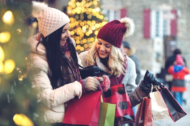 2 ladies smiling and looking at all the shopping bags they are carrying. Ladies are wearing winter coats and hats with puffy beanies.