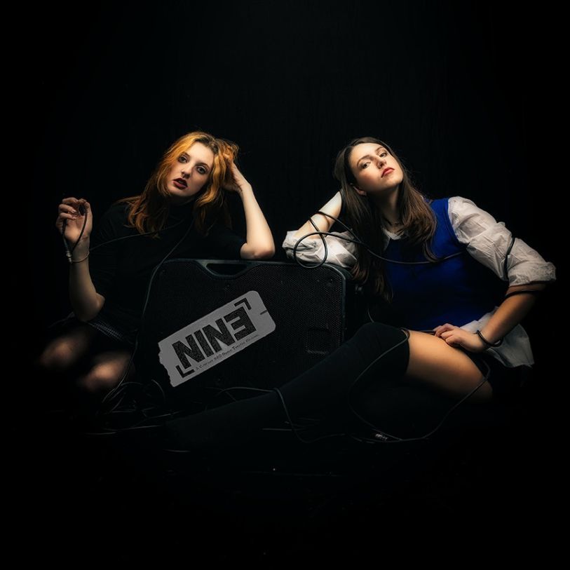 2 girls staring at the camera. The girls are sitting by a big sound speaker with the word NINE on the speaker.