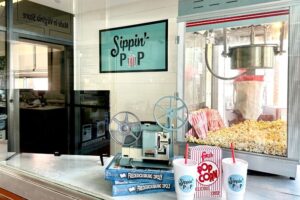 Sippin' Pop front window. Popcorn machine with popcorn on the right. 2 sippin' pop drinks and a box of popcorn in the front.
