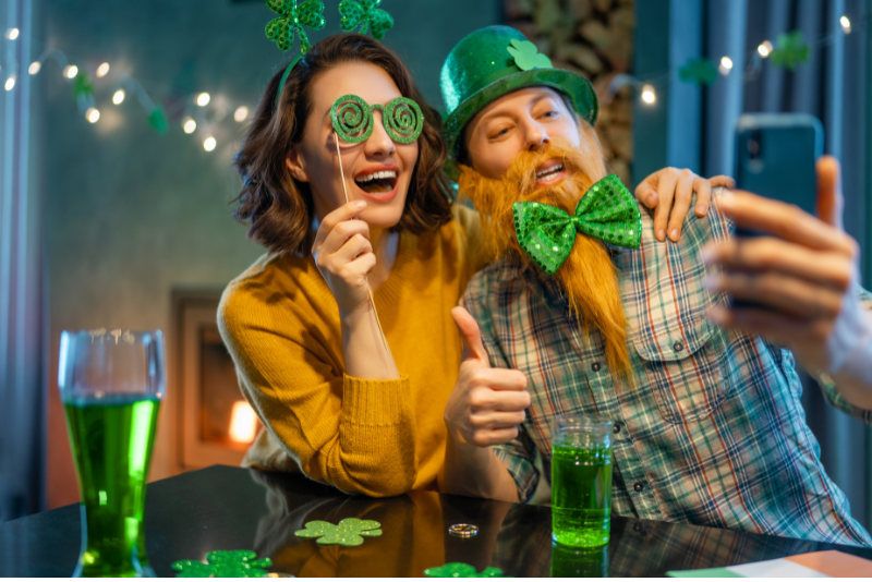 Couple sitting at table with green beer in front of them. Lady on the left has green glitter swirly glasses on. Man on the right has a green top hat and a green bow time. They are smiling at a phone to take a selfie.