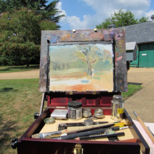 Painting of tree in a wooden frame with paint brushes and paint bottles underneath the painting. Painting supplies are sitting outside with the view of the tree that is being painted.