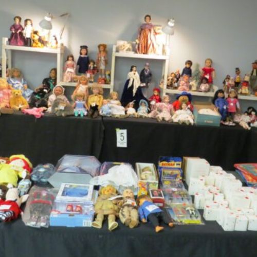 Table at a vendor show with doll and bears on table and on the display behind the table on the wall.