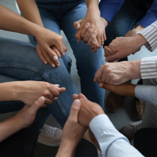 People sitting in a circle holding hands and praying. Images is of only hands and legs.