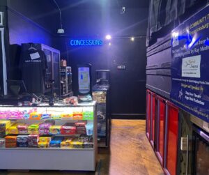 Glass case filled with candy boxes, in front of dark colored concession stand. 
Black wall with a neon blue sign that says 'CONCESSIONS'
Red and black wall to the right. 