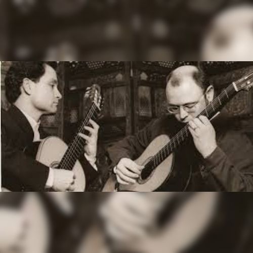 Black and white photograph of 2 men playing guitars. The man on the left in a suite and the man on the right in a dress shirt.