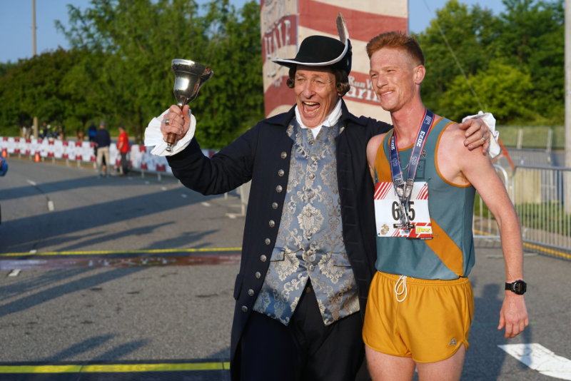 town crier holding a bell in the air with his arm around around a historic half runner. They are standing at the finish line.