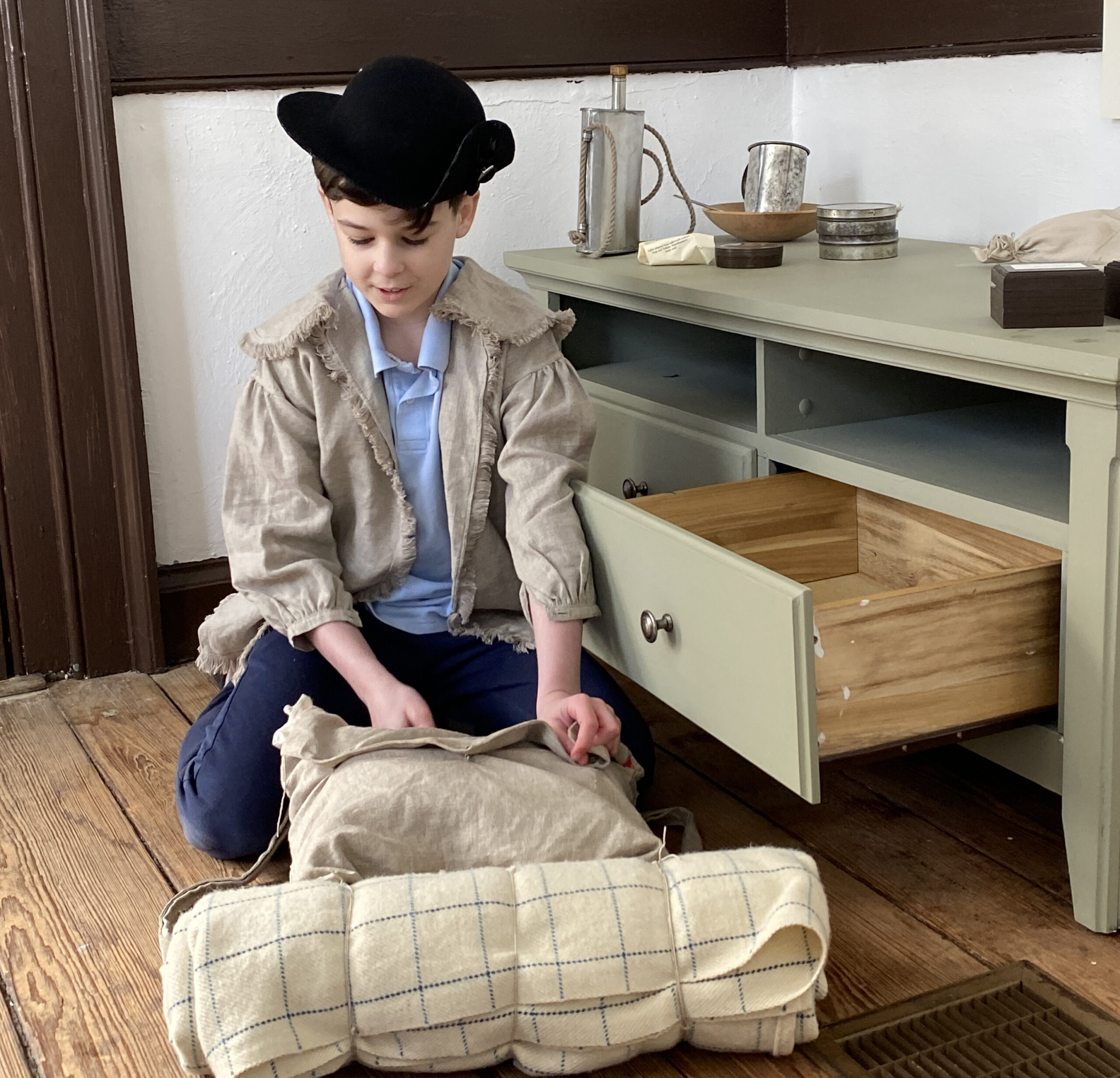boy sitting in front of green dresser with the bottom drawer open. He is looking down at a canvas pillow and an off-white sleeping bag