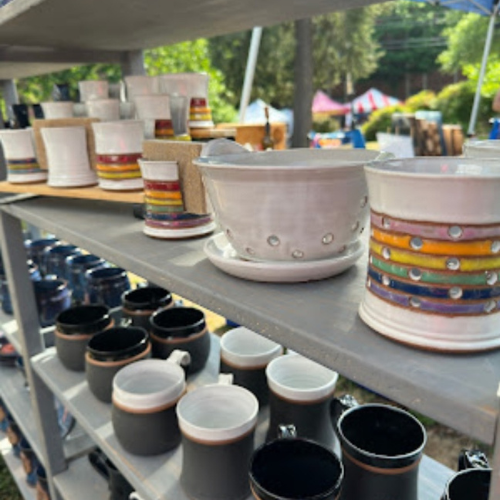 local handmade pottery displayed on shelves outside in the park. Top shelf is white bowls with rainbow bands. Bottom shelf are black mugs.