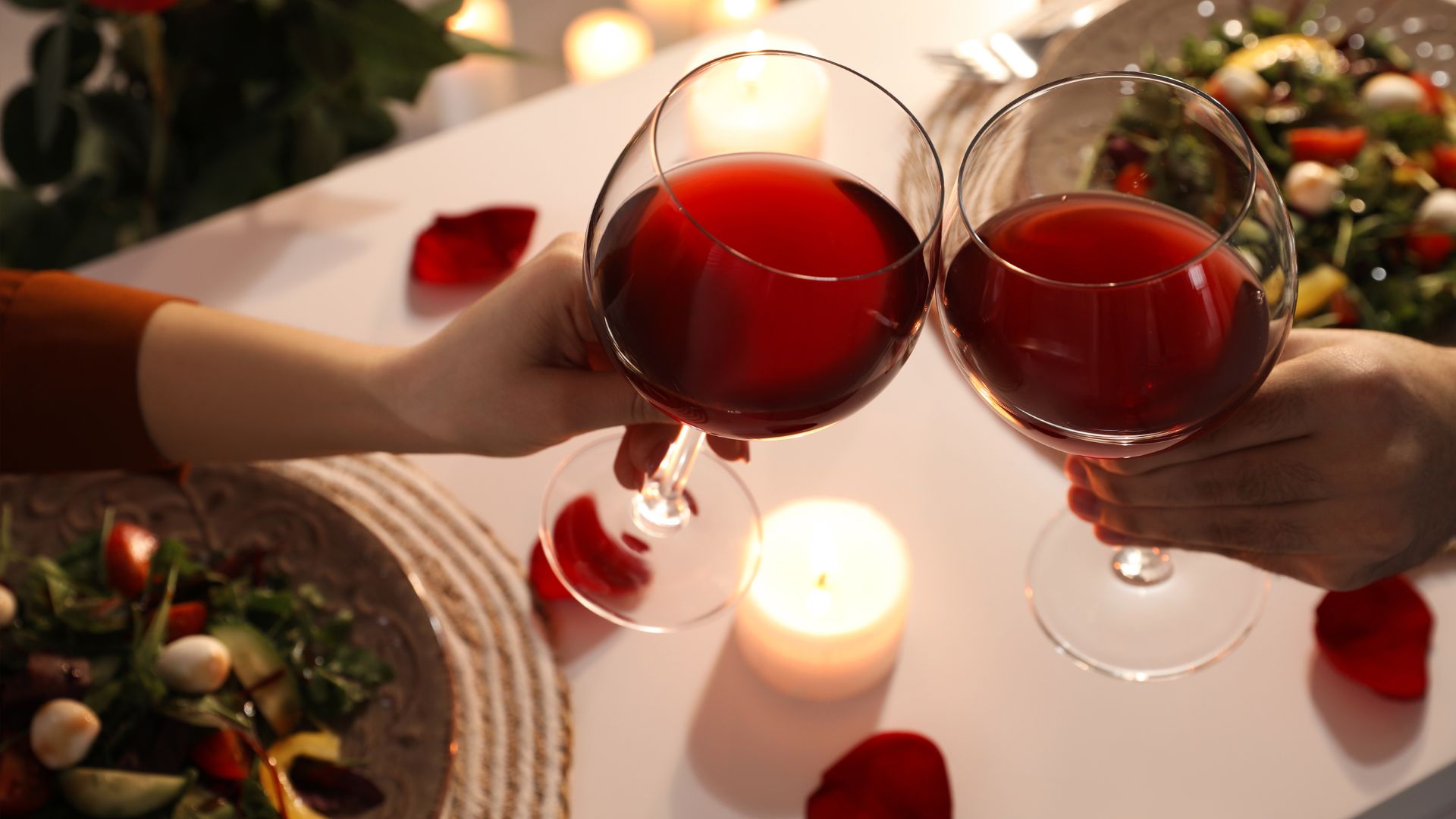 two hands holding glasses of red wine over a dinner table with a candle and rose pebbles