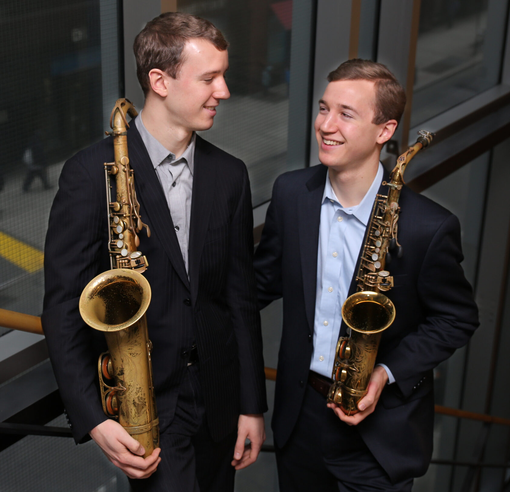 Two young men holding saxophones and smiling at each other.