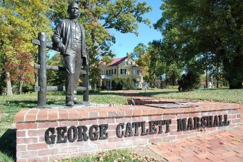 A bronze statue of George Catlett Marshall stands atop a brick wall with his home in the background.