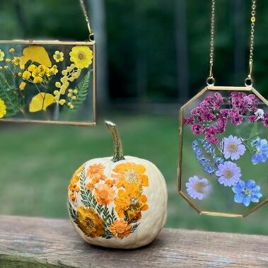 Small painted pumpkin sitting on a wooden bench with two floral glass crafts hanging next to it.