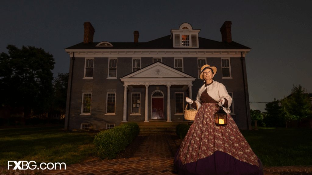 a lady in a Victorian dress holding a lit lantern in front of a brick house giving a ghost tour at dusk/