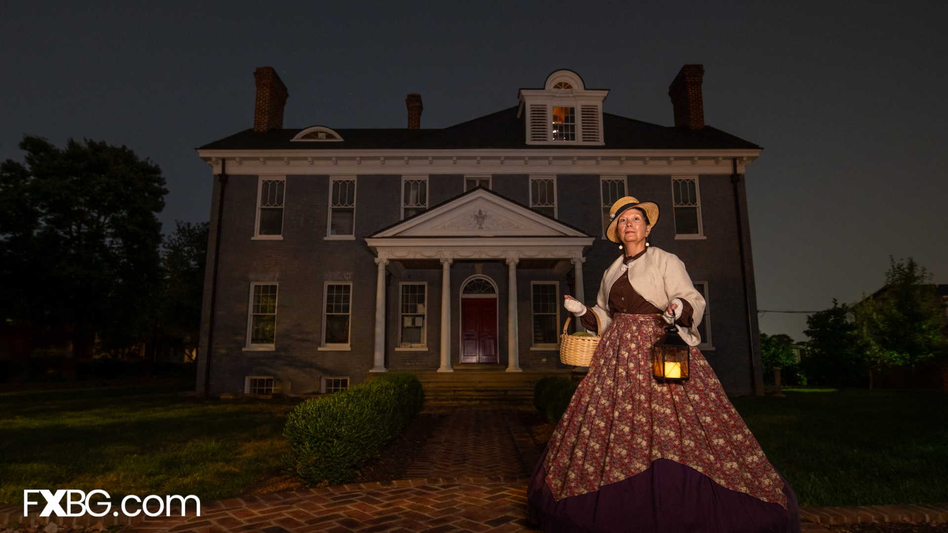 a lady in a Victorian dress holding a lit lantern in front of a brick house giving a ghost tour at dusk/