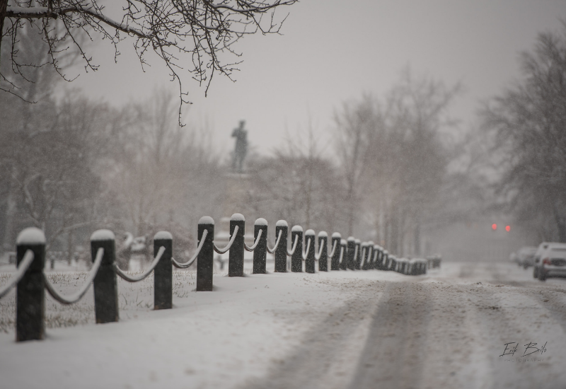 Lots of snow on the road and fence with a statue in the background