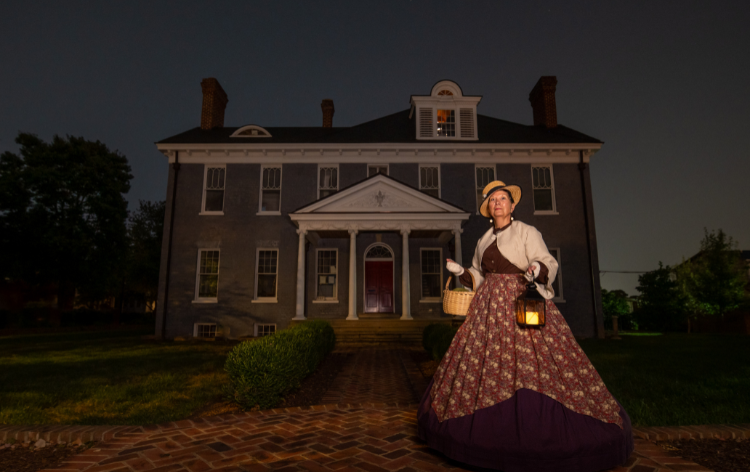 A woman in period dress standing outside of Kenmore holding a lantern