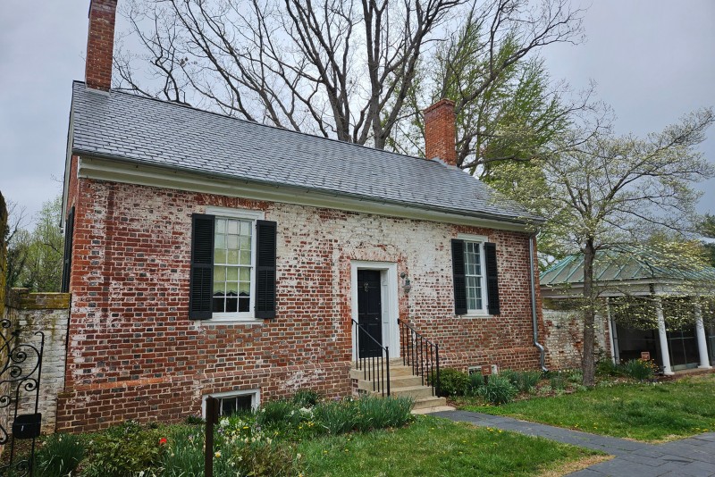 A historic brick building with white washed accents, black shutters, and a slate roof.  A small set of stairs leads to the front door, flanked by windows. A gazebo with a green roof is visible to the right. 