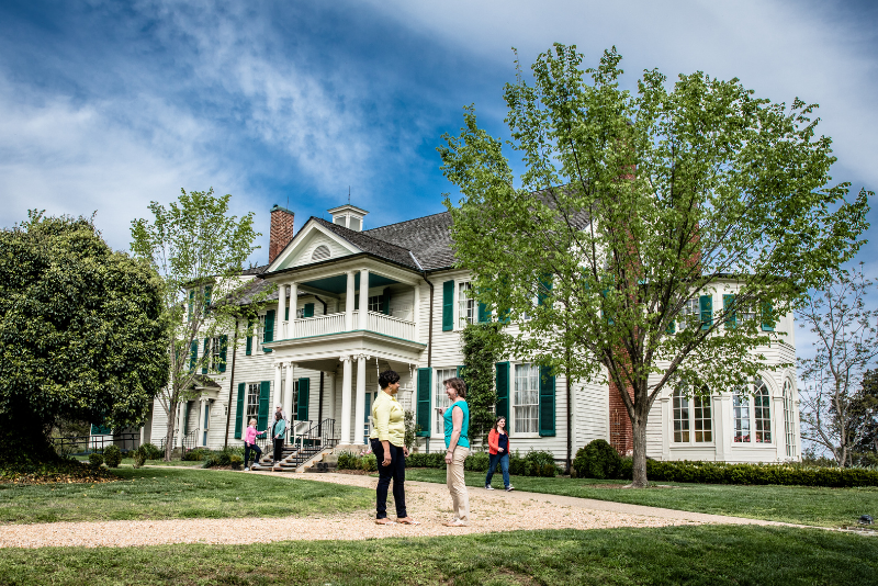 A white colonial style house with green shutters and a wraparound porch stands on a grassy lawn under a blue sky with clouds.  Several people on the grounds, 
