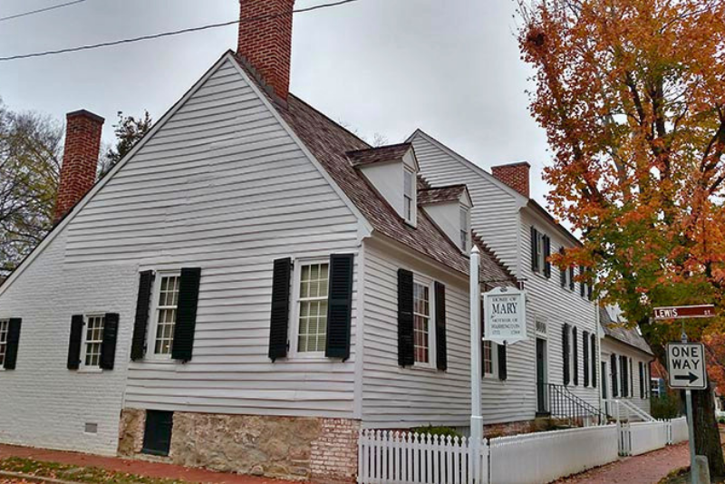 A white clapboard house with black shutters, brick chimneys, and dormer windows, set against a cloudy sky with autumn foliage visible on the right.