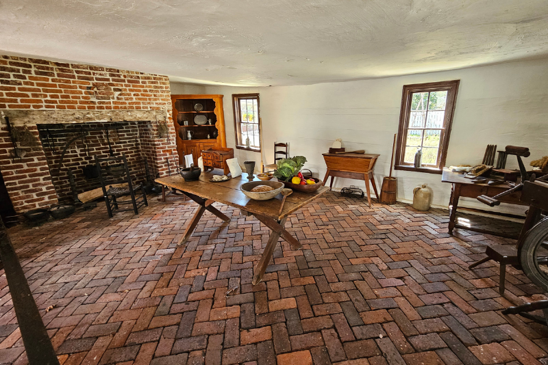 A rustic kitchen with a large brick fireplace, a wooden table laden with produce and a herringbone brick floor. 