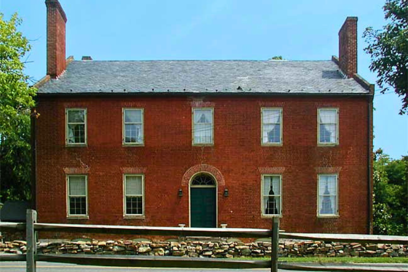 A two-story red brick house with a dark green door and arched brick entryway. They house has a gray slate roof and two prominent brick chimneys.  A low stone wall and wooden fence are in the foreground. 