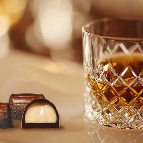 A glass of amber liquid sits next to two chocolates, one of which is cut in half revealing a creamy white filling.