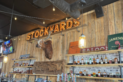 A rustic bar with a mounted bull head, a "STOCKYARDS" sign with marquee lights, shelves of liquor bottles, and a Jameson Irish Whiskey sign.