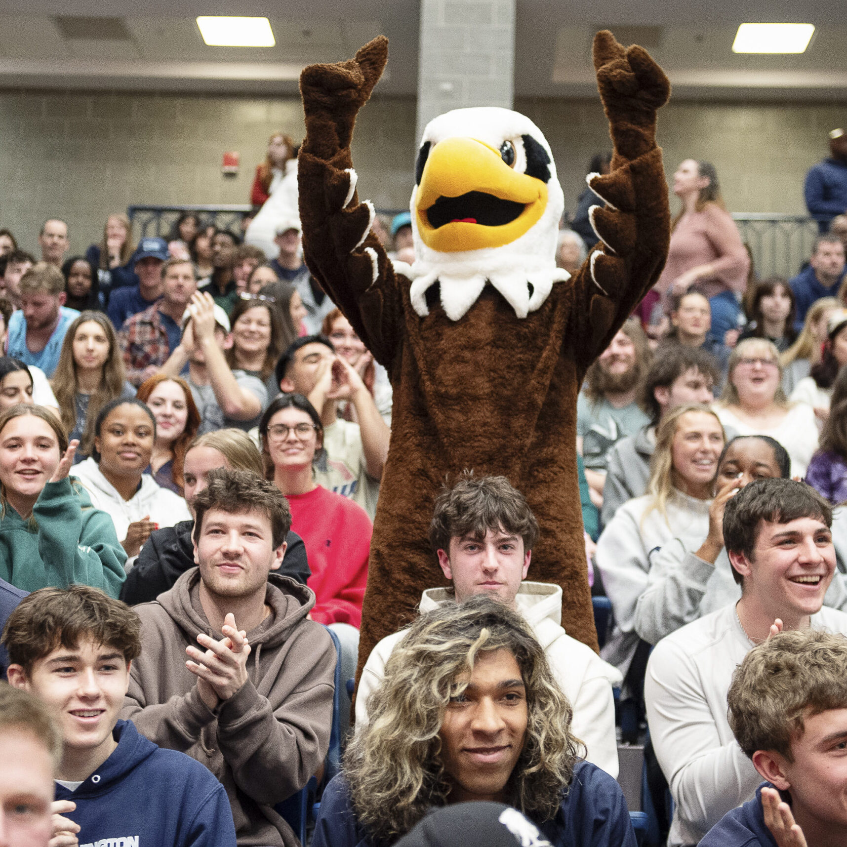 UMW Eagle mascot standing and cheering amongst a crowd of students in the bleachers at a sports game.
