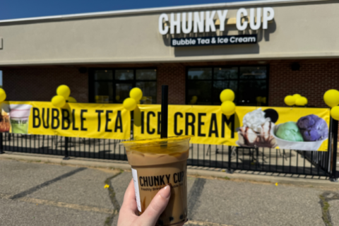 A hand holds a bubble tea in front of a Chunky Cup store, which has a banner advertising "Bubble Tea Ice Cream" and yellow balloons.