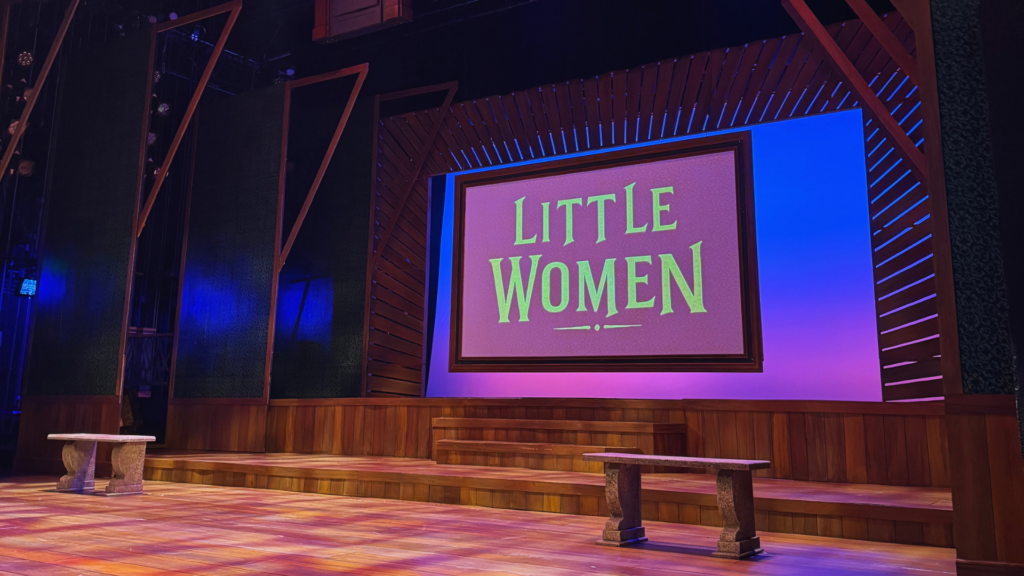 The stage for the musical Little Women features a backdrop with the title in yellow lettering on a pink background, surrounded by wooden beams.