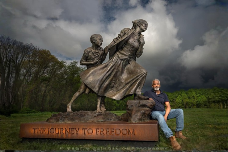 A bronze sculpture titled "The Journey to Freedom" depicts a woman pulling a child along, with a man sitting on the base of the sculpture in front of a wooded background and a stormy sky.