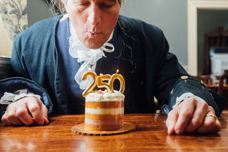 A person in historical attire blows out the candles on a small cake with "250" written on it in gold numbers.