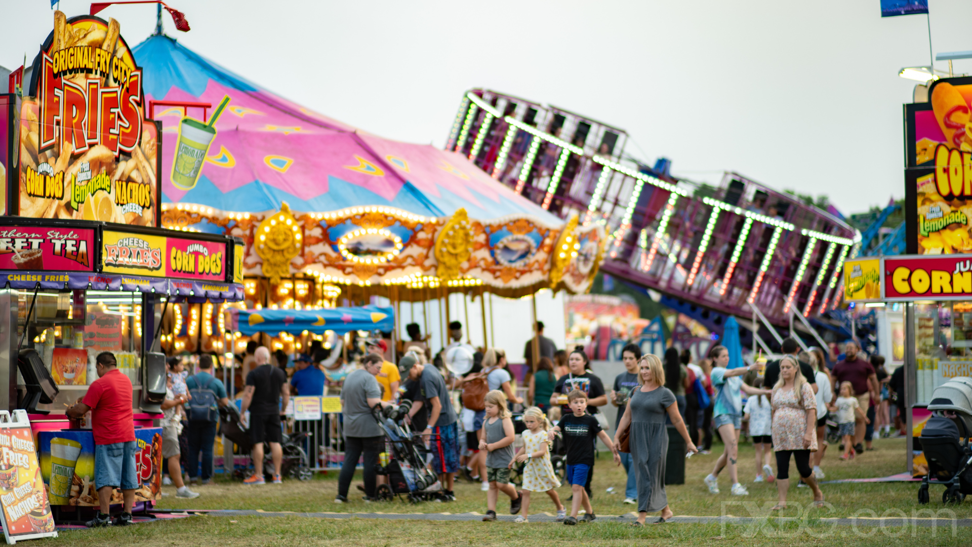 A lively fairground scene with food stalls, rides, and people enjoying themselves under a dusky sky.