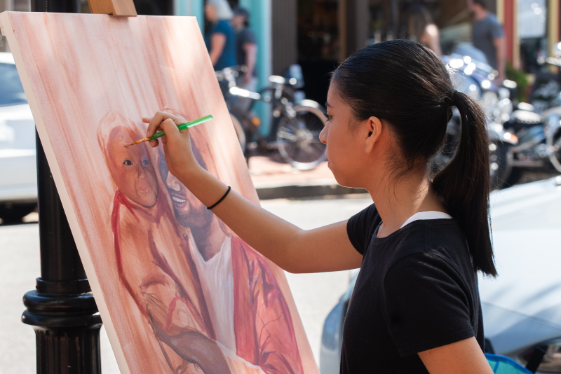 A person painting a portrait on an easel with a green-handled brush, standing at an outdoor work station while bicycles and other people are visible in the background.