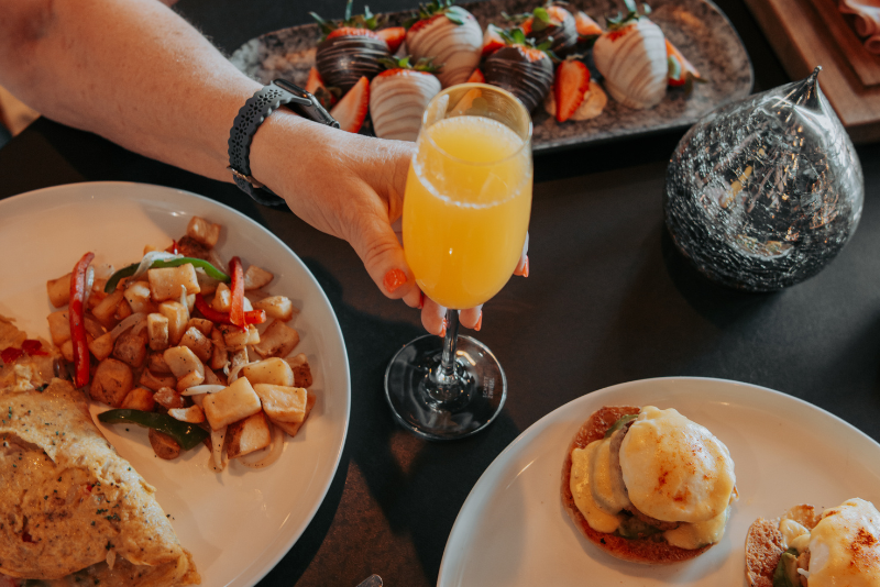 A person is holding a glass of mimosa with chocolate covered strawberries behind the glass. Potatoes and omelet on the left and eggs benedict on the right