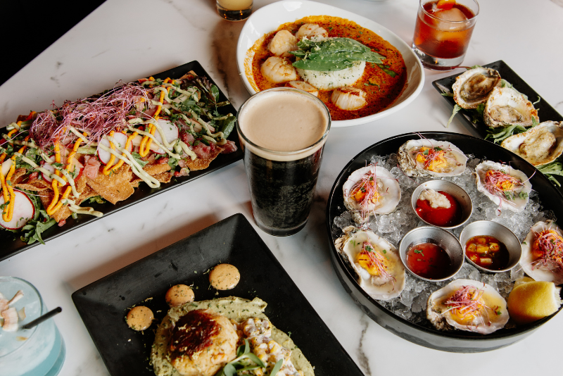 A table set with several dishes: nachos with shredded toppings, a bowl of scallops with green sauce, a plate of raw oysters on ice and sauce, crab cakes and drinks.