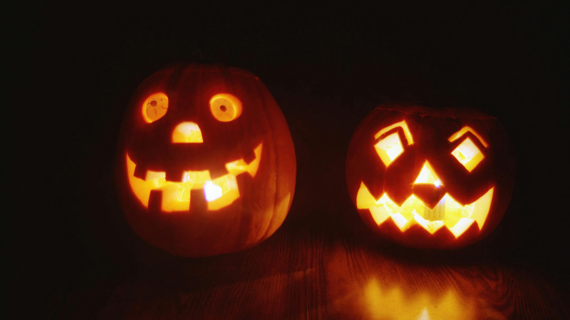 Two carved Halloween pumpkins on a dark wooden surface, left with a glowing jack-o’-lantern face and right with a jagged toothy grin and glowing eyes.