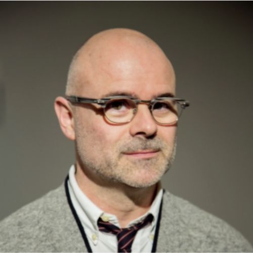 Close-up portrait of a man with a bald head and glasses, wearing a light shirt, patterned bow tie, and gray cardigan against a plain dark background.