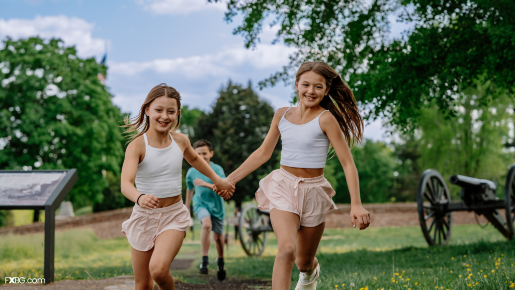 Two children in white tops and light shorts run hand in hand along a park path, with a third child walking behind them, green trees and a cannon in the background.