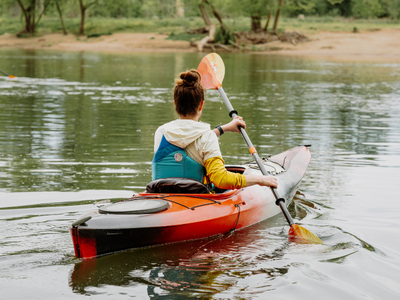 Kayaker paddling a red-and-black kayak on the Rappahannock River, with another person in a canoe: green trees line the riverbank in the background.