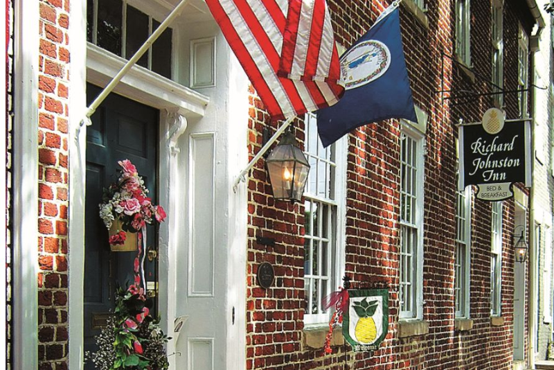 Brick building walkway with a white door, hanging flower baskets, a blue porch banner near the sign “Richard Johnsen Inn,” and striped awning over a small entrance.
