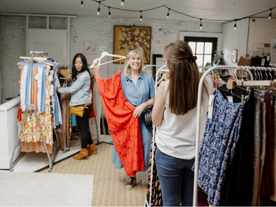 In Wildflower Collective, a woman in a blue shirt holds up an orange dress while a customer browses racks of patterned garments behind her under string lights.